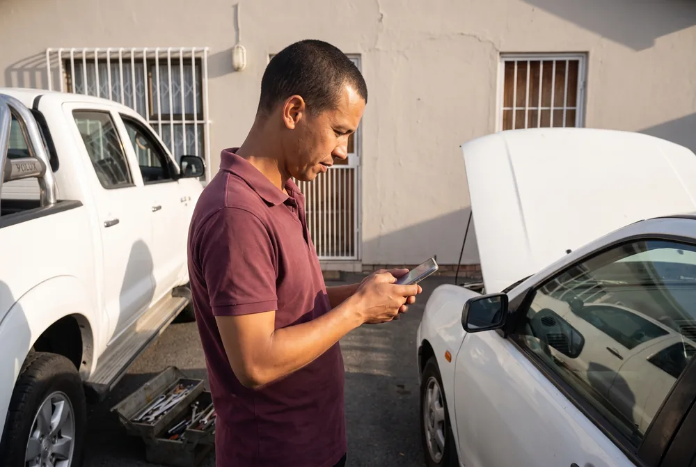 The mechanic checking a customer’s details on his phone in a suburban driveway, his bakkie and open toolbox behind him and the customer’s car with its bonnet up.